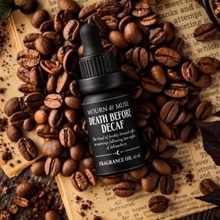A small black dropper bottle labeled 'Mourn & Muse Death Before Decaf Fragrance Oil 10 ml' sits on top of scattered roasted coffee beans over an open book page.