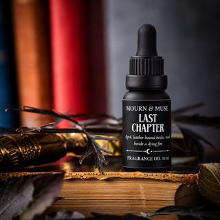 A small black dropper bottle labeled 'Mourn & Muse Last Chapter' fragrance oil stands on an old book, surrounded by dried leaves, a gold-toned object, and blurred books in the background.