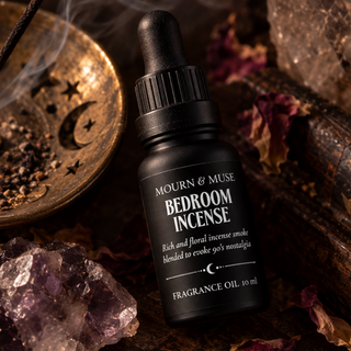 A bottle of Mourn & Muse Bedroom Incense fragrance oil sits on a dark wooden surface surrounded by crystals, dried flowers, and a brass incense dish with smoke rising.