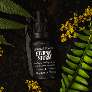 A black dropper bottle of Mourn & Muse Eternal Storm Fragrance Oil, surrounded by green fern leaves and small yellow flowers on a dark, wet background.