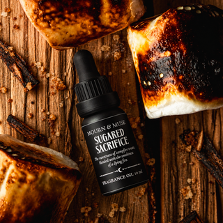 A bottle of Mourn & Muse Sugared Sacrifice fragrance oil lies on a rustic wooden surface surrounded by toasted marshmallows and dark brown spice pieces. The bottle is black with a black dropper cap and white text on the label.